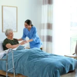 A nurse in blue scrubs hands a glass of water to an elderly woman in bed. The room is bright and airy, with a walker and modern decor, conveying care and comfort.