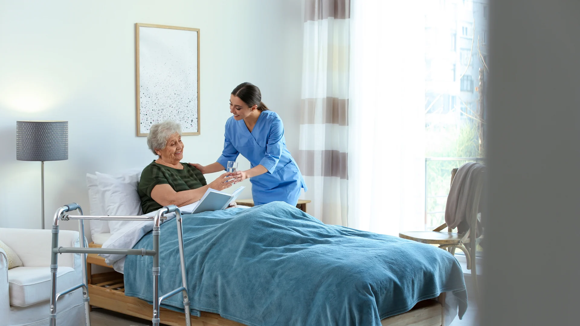 A nurse in blue scrubs hands a glass of water to an elderly woman in bed. The room is bright and airy, with a walker and modern decor, conveying care and comfort.