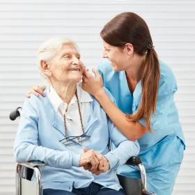 A smiling nurse in blue scrubs gently touches an elderly woman's face who is seated in a wheelchair, both appearing joyful and connected.