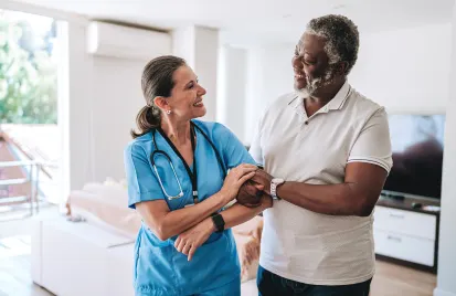 A smiling nurse in blue scrubs warmly holds the arm of a happy older man in a white shirt. They share a moment in a bright, cozy living room.