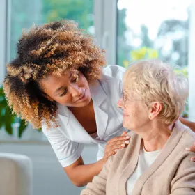 A caregiver with curly hair smiles warmly at an elderly woman wearing glasses and a cardigan. They're indoors, fostering a caring and supportive atmosphere.