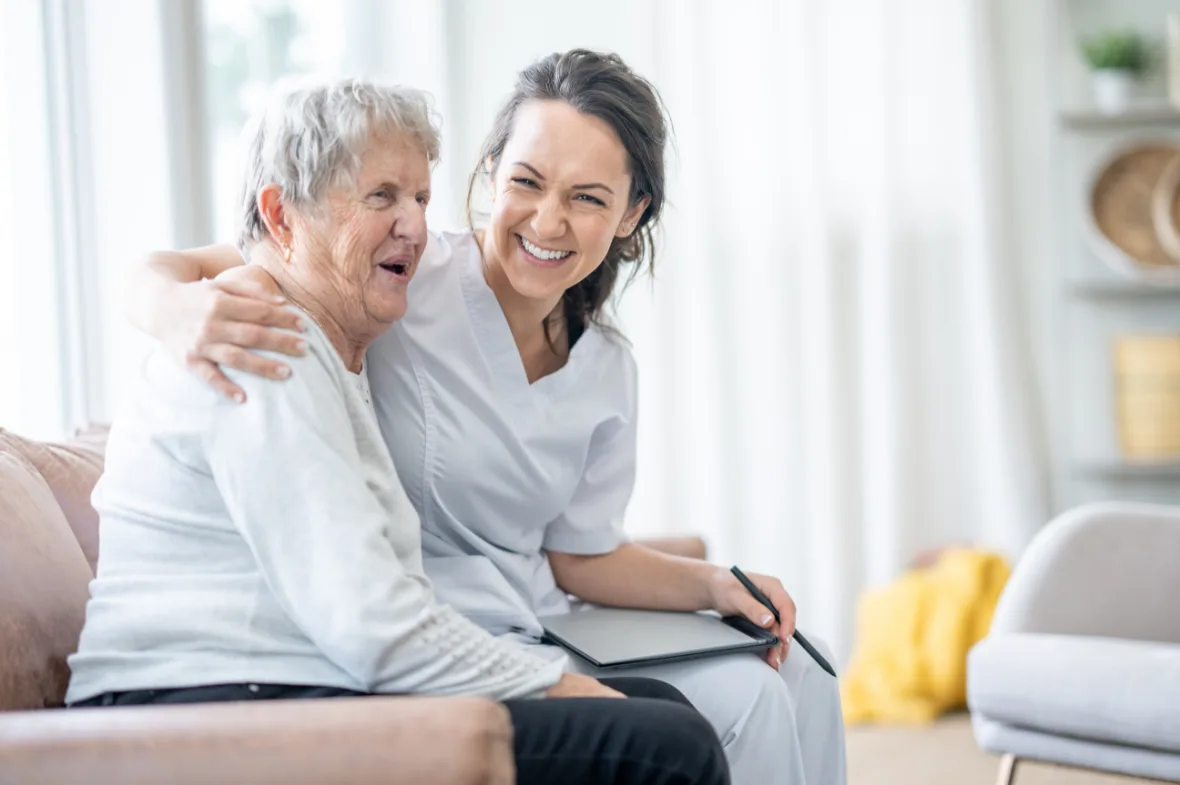 A caregiver in white scrubs warmly embraces an elderly woman on a couch, both smiling joyfully. The atmosphere is bright and comforting.