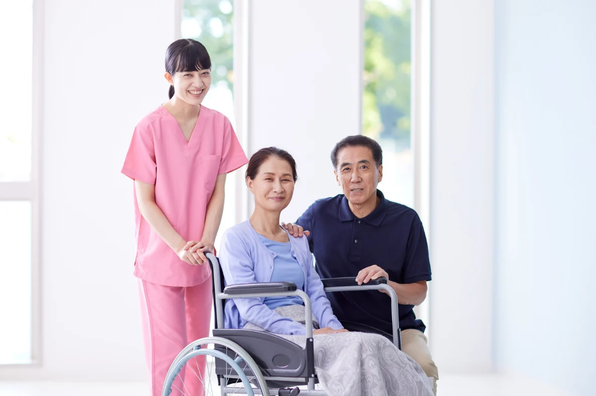 A smiling nurse in pink scrubs stands beside a woman in a wheelchair, accompanied by a man. The setting is bright, conveying a caring and supportive atmosphere.
