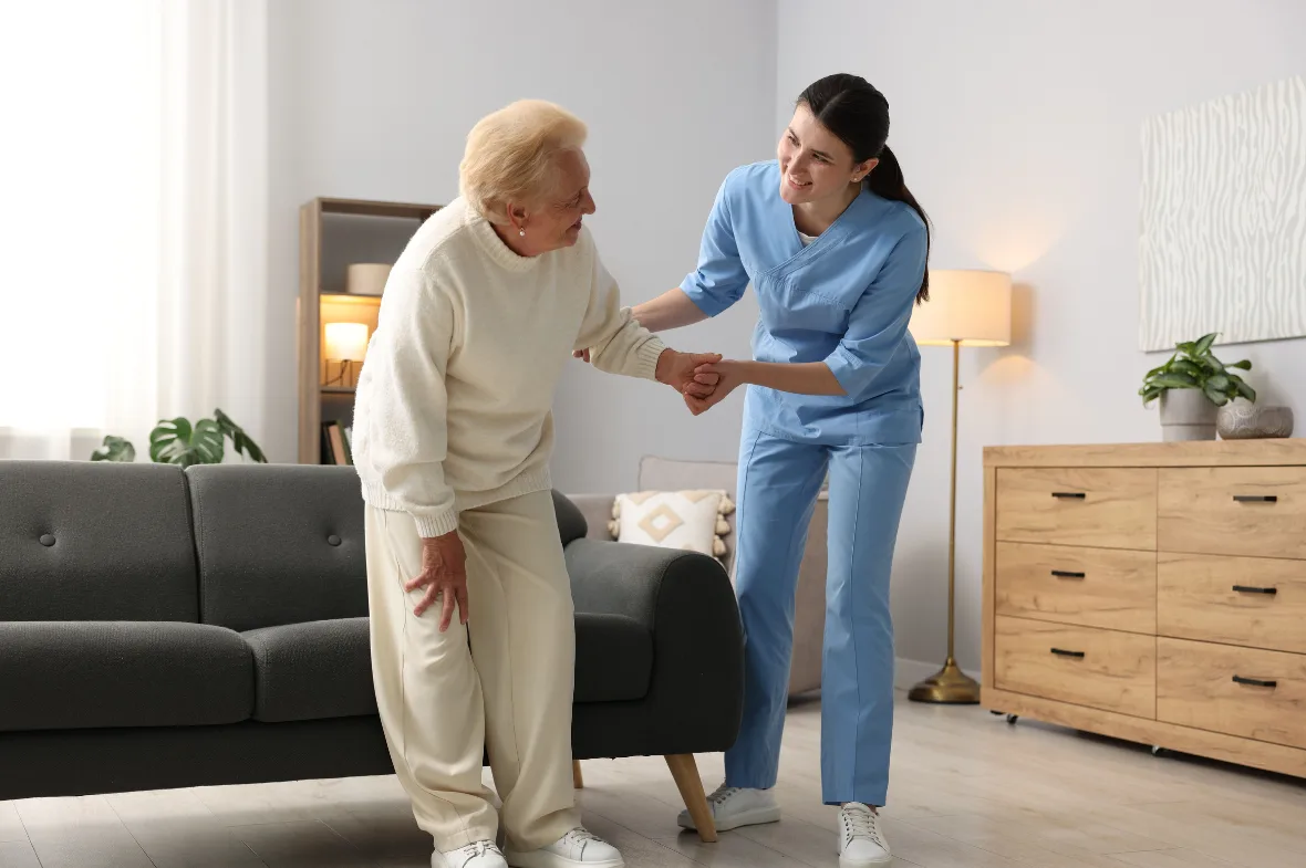 A caregiver in a blue cardigan hands a cup to a smiling elderly woman seated in a cozy armchair, conveying warmth and care in a bright living room.