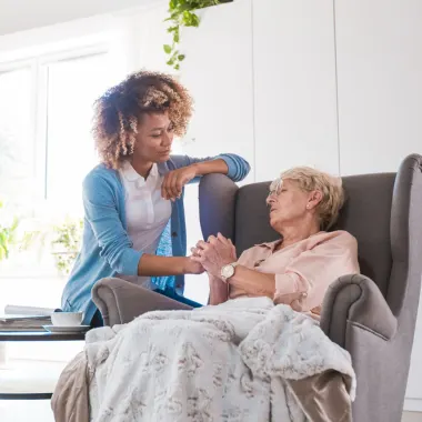 A young woman tenderly holds an older woman's hand as she sits in an armchair, conveying care and empathy in a bright, airy room.