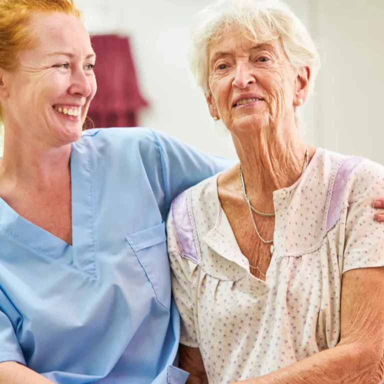 A caregiver in blue scrubs sits beside a smiling elderly woman in a nightgown, sharing an affectionate moment. The room is bright and cozy.