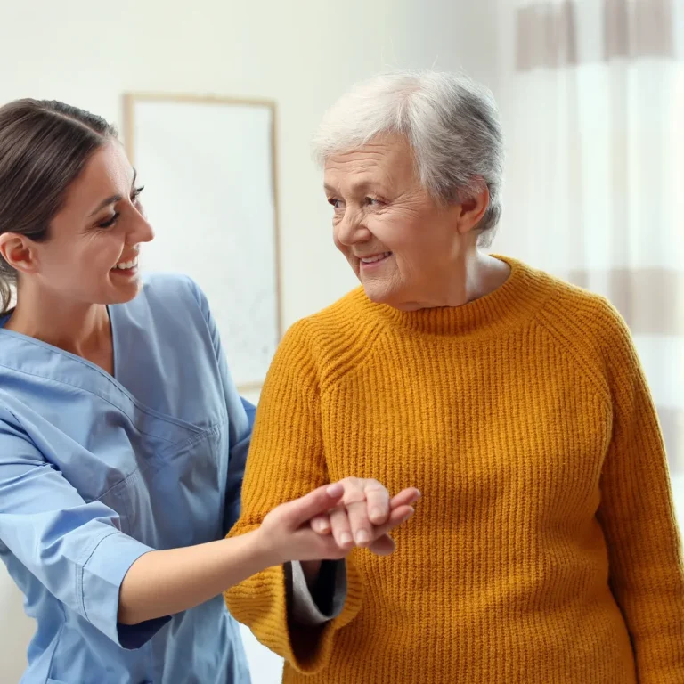 A nurse in blue scrubs warmly assists an elderly woman in a cozy yellow sweater. They share a joyful moment in a well-lit room, conveying care and support.